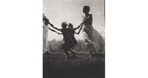 A black-and-white photograph of a White woman and children wearing long dresses holding hands to form a circle in a grassy field.