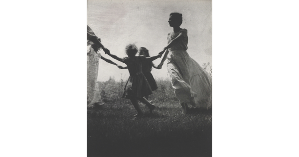 A black-and-white photograph of a White woman and children wearing long dresses holding hands to form a circle in a grassy field.
