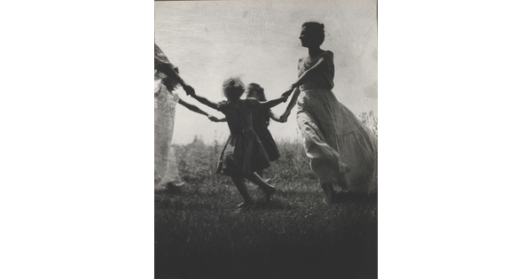 A black-and-white photograph of a White woman and children wearing long dresses holding hands to form a circle in a grassy field.