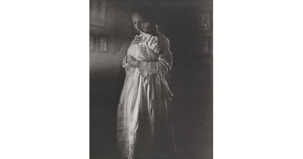 A black-and-white photograph of a standing White woman holding a baby wearing a long white nightgown.