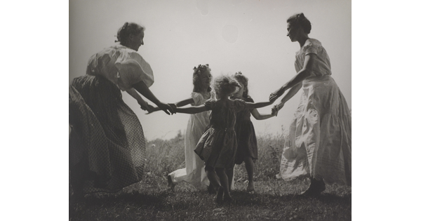 A black-and-white photograph of White women and children wearing long dresses holding hands to form a circle in a grassy field.