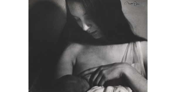 A black-and-white photograph of a White woman with long, dark hair breastfeeding a baby.