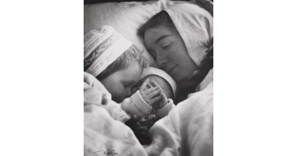 A black-and-white photograph of a White woman in a nightcap cuddling in bed with a toddler and an infant.
