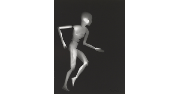 A black-and-white photograph of a white person-shaped puppet running on a black background.