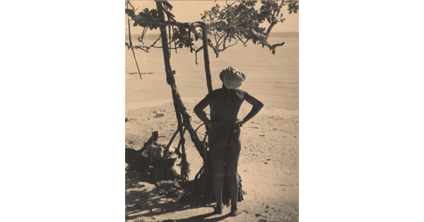 A sepia-toned photograph of a nude dark-skinned woman, hands on hips, with a swath of fabric wrapped around her head looking up at a tree.