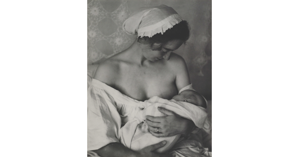 A black-and-white photograph of a White woman in a nightcap and white flowing blouse nursing an infant.