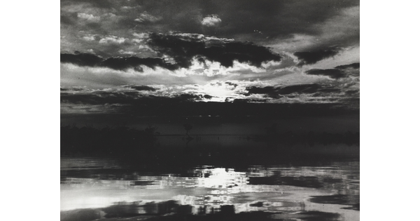 A black-and-white photograph of a cloudy sky with sun peeking through reflected on water below.