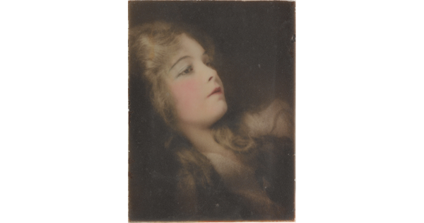 A hand-colored, cropped photograph of a reclining woman with long hair and flushed pink cheeks.