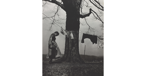 A black-and-white photograph of a White woman in a dress and apron standing next to a large tree with a clothesline and clothes hanging from it.
