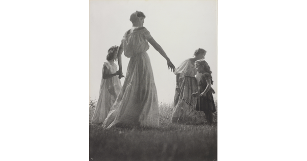 A black-and-white photograph of White women and children wearing long dresses holding hands to form a circle in a grassy field.