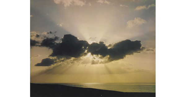 A color photograph of a sun peeking from behind clouds over a calm beach.