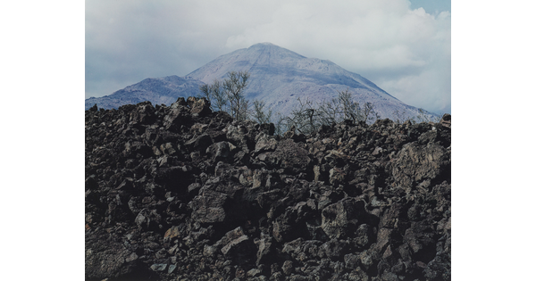 A color photograph of a hill of black stones, the tops of some evergreens, and a mountain peak in the background.