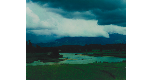 A color photograph of storm clouds rolling over a mountain with a winding river in the foreground.