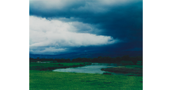 A color photograph of storm clouds rolling over a mountain with a winding river in the foreground.