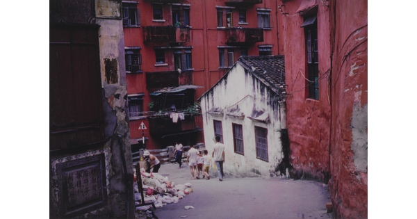 A color photograph of people walking down a garbage-strewn sloping street in front of tall red buildings and a single-story white building.