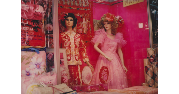 A color photograph of two White Barbie-like dolls, one wearing an Asian-style red and gold outfit and the other a pink gown, in a shop window.