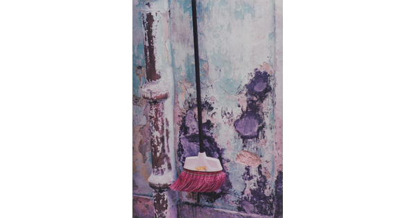 A color photograph of a dark pink bristle broom hanging on a plaster wall with light blue peeling paint.