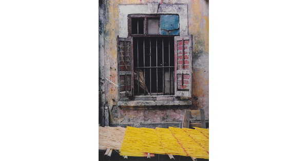 A color photograph of a barred window, wood shutters open, set in peeling yellow plaster above a yellow thatched awning.