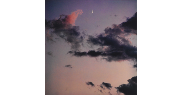 A color photograph of a crescent moon at dusk above thin, dark clouds.