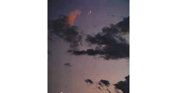 A color photograph of a crescent moon at dusk above thin, dark clouds.