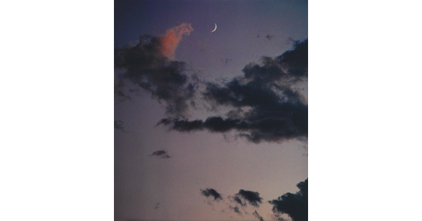 A color photograph of a crescent moon at dusk above thin, dark clouds.