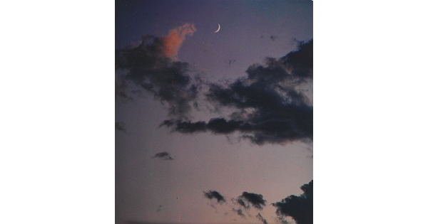 A color photograph of a crescent moon at dusk above thin, dark clouds.
