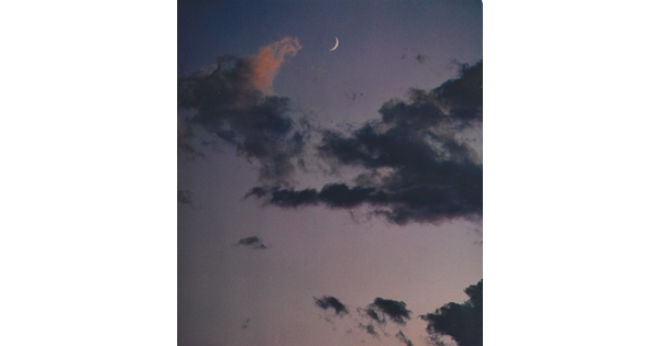 A color photograph of a crescent moon at dusk above thin, dark clouds.