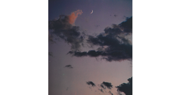 A color photograph of a crescent moon at dusk above thin, dark clouds.