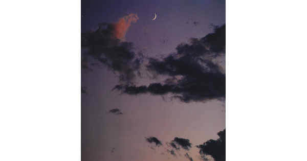 A color photograph of a crescent moon at dusk above thin, dark clouds.