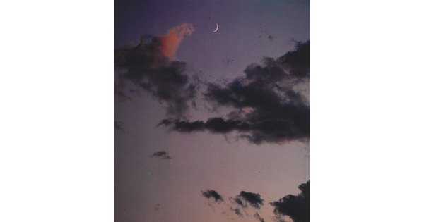 A color photograph of a crescent moon at dusk above thin, dark clouds.