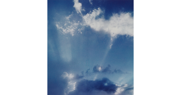 A color photograph of white clouds in a blue sky and sun rays that create white streaks.