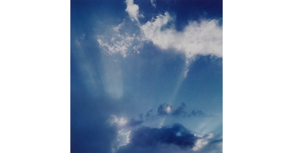 A color photograph of white clouds in a blue sky and sun rays that create white streaks.