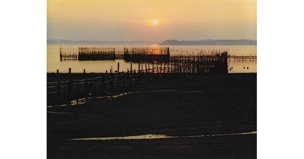 A color photograph of a sunset over a bay with a rocky beach and a wooden dock in the foreground.
