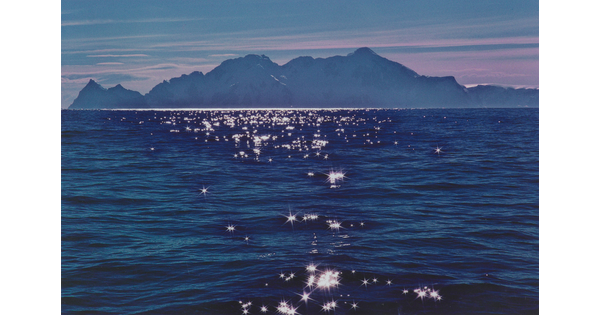 A color photograph of a dark mountain range under a pink sky beyond gentle bright blue waves with light reflecting off of the ripples.