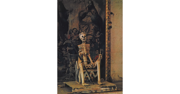 A color photograph of a skeleton seated on a throne, wearing a crown and holding a scythe in one hand, in front of a painting.