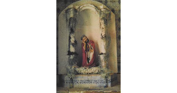 A color photograph of a painted sculpture of a seated Jesus wearing a crown, red robe, holding a cane, head resting on his hand in an arched niche.