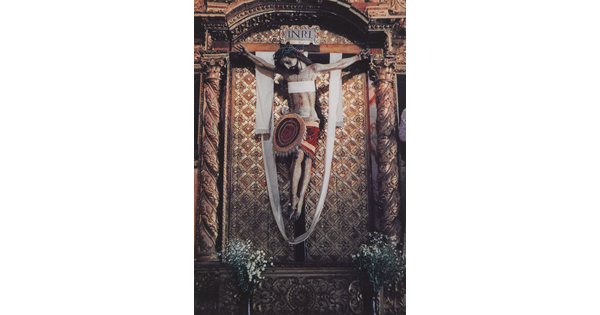 A color photograph of a painted sculpture of a crucified Jesus wearing a crown of thorns, a white cloth draped over the cross, hanging on an ornate gold wall.