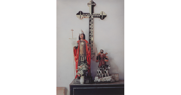 A color photograph of a table with figures of a saint in a red robe holding a cross staff, and a cowherd with two cows; a large black-and-white cross in the background.