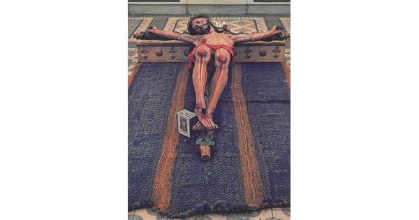 A color photograph of a painted sculpture of a crucified Jesus, eyes closed, wearing a crown of thorns; the entire crucifix laying on a blue cloth on a tile floor.