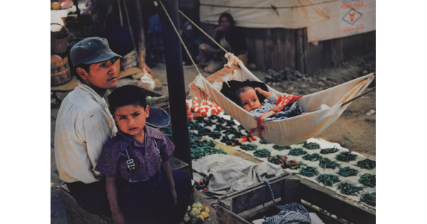 A color photograph of a medium-skinned man, child, and baby; the man seated wearing a cap, child standing next to him as the baby lies in a hammock in front of them.