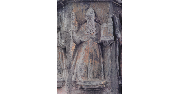 A color photograph of a stone figure with a long beard, wearing robes, holding a building in one hand and what might be a staff in the other.