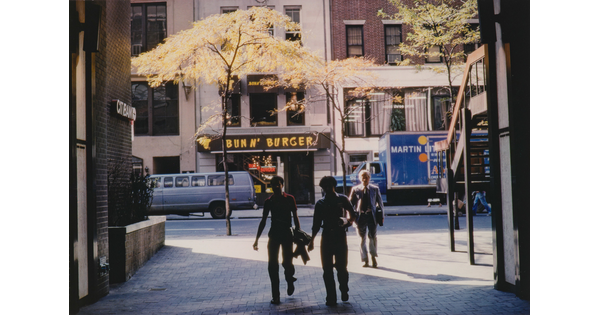 A color photograph of three men walking in an alley paved with bricks; behind them are small trees lining the sidewalk and a restaurant on the opposite side of the street.