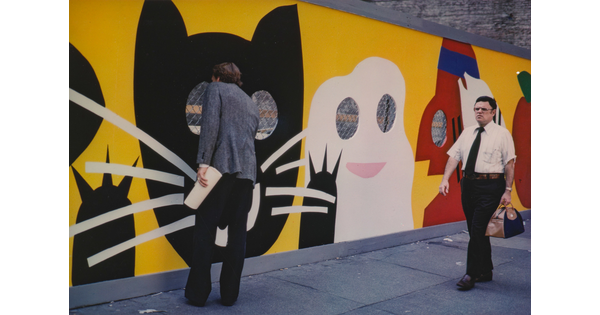 A color photograph of two men walking past a construction fence decorated with a cartoonish black cat, ghost, and other characters; one man peeks through the fence.