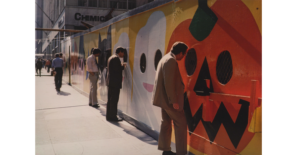 A color photograph of men in business attire on a city sidewalk looking through holes in a construction wall that is painted with a mural of pumpkins and ghosts.