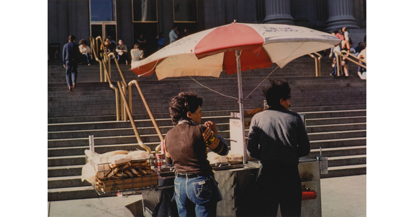 A color photograph of a food vendor at a cart on the sidewalk as people go up and down steps leading to a building with fluted pillars.