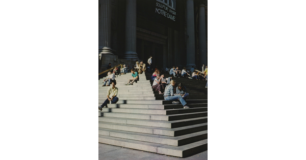 A color photograph of people sitting on the stone steps of a building with large fluted columns.