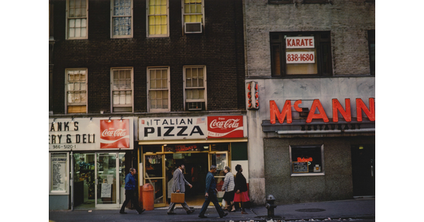 A color photograph of pedestrians walking past storefronts advertising "Italian Pizza" and "Coca-Cola."