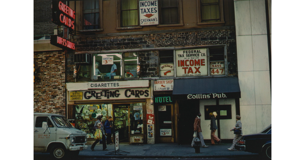 A color photograph of the front of a building advertising a greeting card shop, a hotel, a pub, and other services as people walk past.