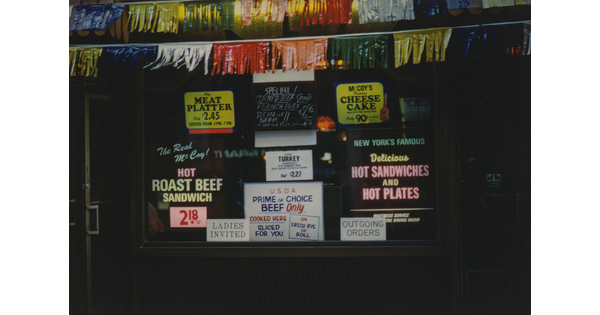 A color photograph of shop window, framed by colored fringed garlands, covered in advertisements for various foods for sale.
