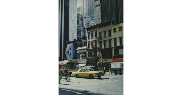 A color photograph of a busy city street with pedestrians, cyclists, cars, and busses.